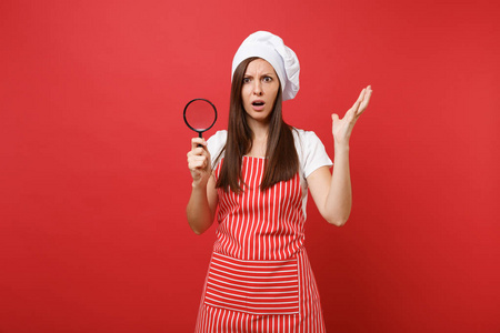 家庭主妇女厨师或面包师穿条纹围裙，白色T恤，烤厨师帽子隔离在红墙背景上。 女管家透过放大镜看着。 模拟复制空间概念