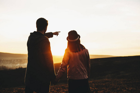 站在田野里，年轻的男人和女人牵着手欣赏壮丽的日落景色。无法辨认的一对看日落的夫妇