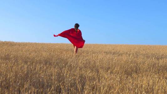 穿着红色雨衣的年轻女孩扮演超级英雄，穿过黄色小麦的田野，对抗蓝天
