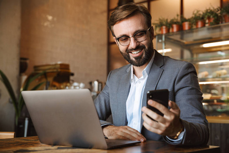 一位年轻英俊的年轻商人，穿着西装，在一台笔记本电脑上工作，坐在咖啡馆里拿着手机