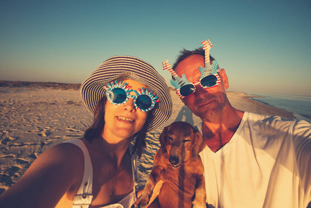 欢快的夫妇戴着有趣的太阳镜，带着一只小狗在海滩上自拍，在日落时在蓝天的背景下玩得很开心。 自然界中快乐的人。 复古的形象。