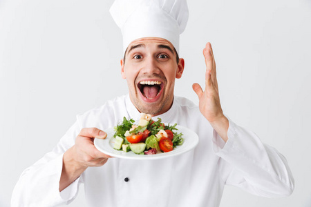 令人兴奋的男厨师穿着制服，在白色背景的盘子上展示新鲜的绿色沙拉