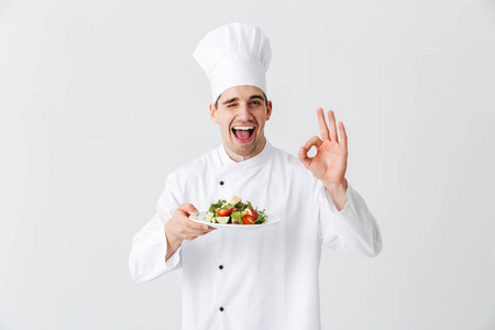 兴奋的男人厨师穿着制服，在白色背景的盘子上展示新鲜的绿色沙拉，好吗