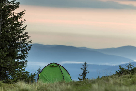 在遥远的雾蒙蒙的蓝山上的草山上的旅游帐篷，日出或日落背景前清澈的粉红色天空。 黎明时在山上露营。 旅游徒步旅行和自然美景的概念。
