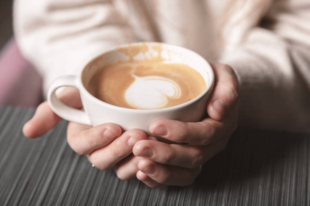 关上一杯白色的热拿铁艺术咖啡，一个年轻女孩手中有一个心形。 女人穿着温暖的冬季针织白色毛衣。 变调了。 咖啡女孩概念放松咖啡杯。