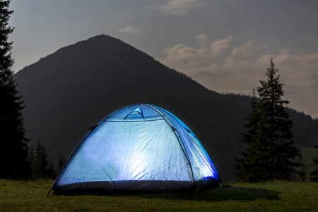 旅游徒步旅行者的帐篷在绿绿的山谷里，在深蓝色的多云天空下，在覆盖着森林背景的远山上，明亮地从里面点燃。夏天晚上在山上露营。