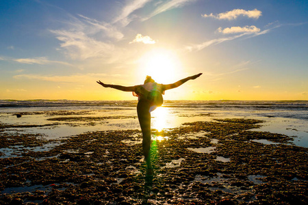 年轻健康的女人在日落时在海滩上练习瑜伽。 在海边夕阳下坚强的自信女人。 年轻女人在海滩上做瑜伽的剪影。
