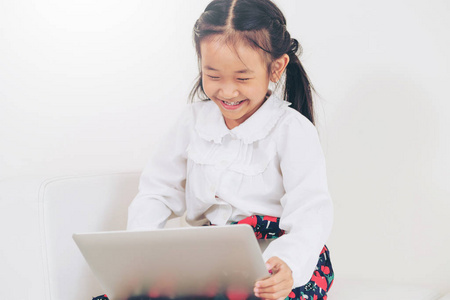 小快乐的孩子用笔记本电脑坐在白色沙发上。 童年生活方式。