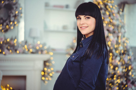 怀孕爱人和期望的概念幸福的孕妇，穿着牛仔裤站在圣诞树的背景下的房间形象
