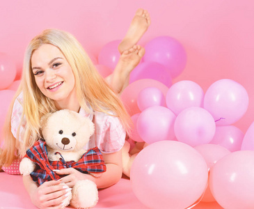 生日女孩的概念。女孩在睡衣, 家居服躺在气球附近, 粉红色的背景。金发碧眼的笑脸放松与泰迪熊玩具。妇女可爱庆祝生日与气球