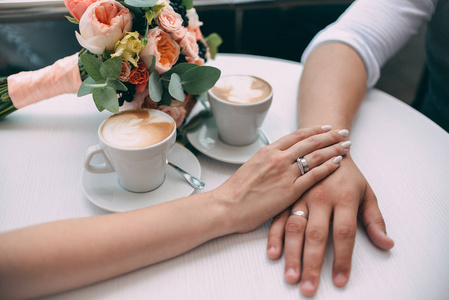 新郎新娘手牵着手, 拿着昂贵的彩礼戒指和白金, 背景是婚礼花束和白色桌子上的两杯咖啡