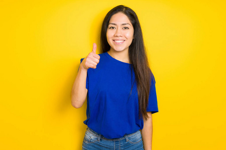 美丽的黑发女人穿着蓝色的T恤，在黄色的孤立的背景下，用手做快乐的竖起大拇指的手势。 赞许的表情看着相机，显示出成功。