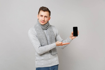 穿着灰色毛衣围巾的年轻人用手指着手机，灰色背景上隔离着空白的空屏幕。 健康的生活方式，人们真诚的情感，寒冷的季节概念。 模拟复制