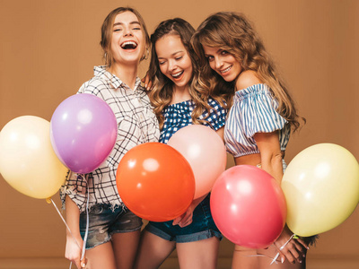 三个微笑的漂亮女人穿着格子衬衫夏天的衣服。 在金色背景下摆姿势的女孩。带彩色气球的模特。准备庆祝生日或节日聚会的乐趣