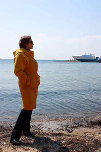 一个年轻的女人穿着黄色的外套站在海滩上。 见过游艇。