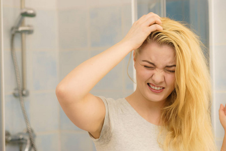 金发女人有问题，油腻油腻的头发看着自己在浴室。 女性显示头皮划伤自己头皮屑问题。