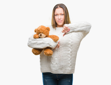 年轻的白种人女人抱着泰迪熊，在孤立的背景下愤怒地面对消极的迹象，表示不喜欢，竖起大拇指拒绝概念