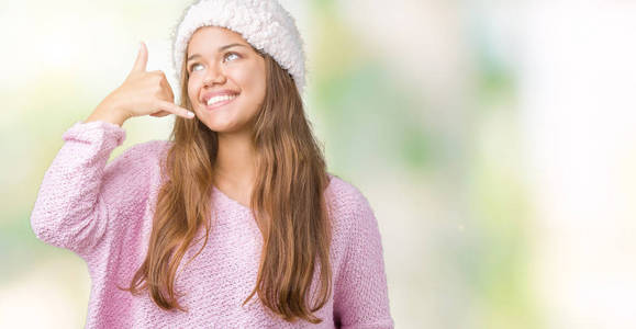 年轻漂亮的黑发女人，穿着毛衣和冬帽，在孤立的背景上微笑，用手和手指做电话手势，就像在电话里说话一样。 交流概念。
