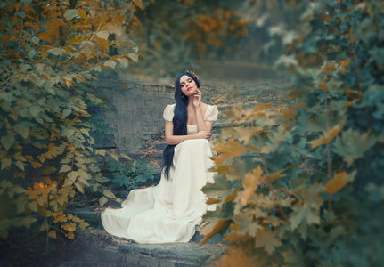 希腊女神坐在秋林的石阶上, 皮肤轻盈, 白色长典雅的礼服, 像雕塑, 黑发与金色桂冠花环, 宏伟的公主新娘婚礼日
