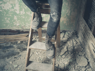 一个年轻人的腿坐在一个凌乱而废弃的房间里的梯子上