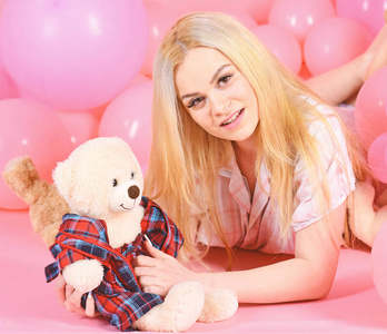 金发女郎在微笑的脸上放松与泰迪熊玩具。女人逗人喜爱用气球庆祝生日。穿睡衣的女孩, 国内的衣服放在空气气球附近, 粉红色的背景。生