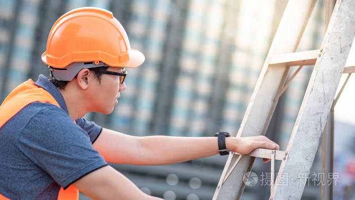年轻的亚洲维修工人,带橙色安全帽和背心,在施工现场携带铝步梯.