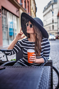 一个年轻的时尚女人戴着太阳镜, 戴着时髦的帽子, 坐在夏季街头咖啡馆里拿着一杯咖啡的肖像