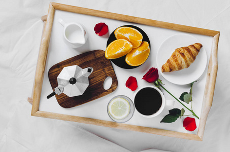 浪漫的情人节早餐在床盘，牛角面包杯咖啡浓缩咖啡，牛奶，新鲜橙子，一杯水，柠檬和红玫瑰。 早上好，概念。 享受缓慢的生活。 上面的