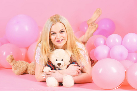 穿睡衣的女孩, 国内的衣服放在空气气球附近, 粉红色的背景。金发女郎在微笑的脸上放松与泰迪熊玩具。女人逗人喜爱用气球庆祝生日。生