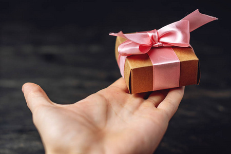 手拿节日礼品盒包装在工艺纸与粉红色丝带在深色木制背景。 精美礼品贺卡的概念