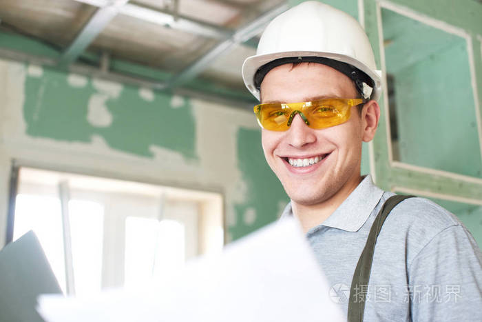 戴着头盔和眼镜的年轻建筑工人微笑着站在一个建筑工地的背景下,手里