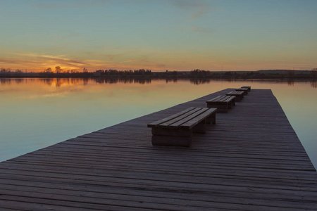 一个宽阔而长的木制码头，在平静的湖面上有日落的长凳