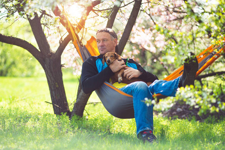 快乐的人和他的有趣的狗达克顺德放松在一个吊床上的绿色草地上，在一个野生盛开的樱桃享受生活，在一个可怕的春天，与自然和谐。 背光。