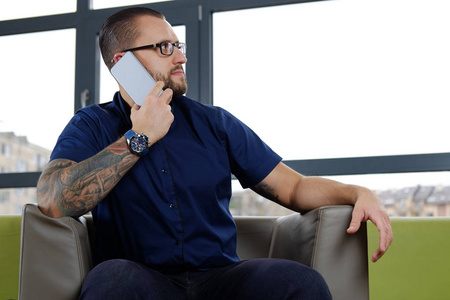 纹身胡须的商人穿着一件蓝色衬衫, 坐在办公室的沙发上, 通过电话交谈