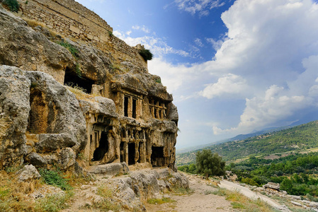 土耳其山区的古代遗迹，左边是一座大山，山上雕刻着房间和柱子，右边是山和树的景色