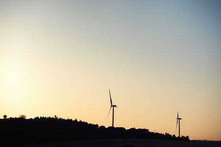 日落时在风电场中产生清洁能源的两个风车的轮廓。 他们在一个被庄稼和树木包围的农村环境中，天空是干净和清澈的。 复制空间