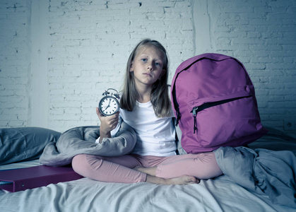美丽的金发小女孩悲伤的失眠和愤怒，显示闹钟时间为上学做好准备，在早上醒来时，孩子们失眠和睡眠障碍的概念。
