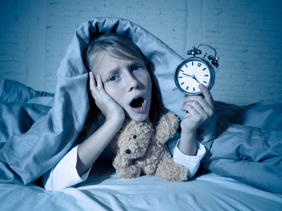 可爱的失眠小女孩躺在床上，闹钟看起来很累，有睡眠问题，晚上睡觉，或者在孩子失眠焦虑和睡眠障碍时醒得太早。