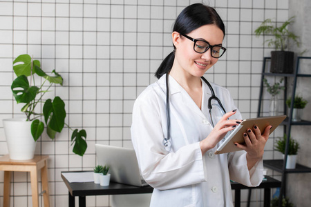 女性黑发医生戴眼镜使用平板电脑, 聪明可爱的女人与小工具和听诊器在白色的办公室背景