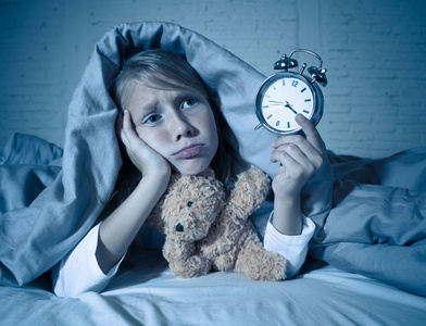 可爱的失眠小女孩躺在床上，闹钟看起来很累，有睡眠问题，晚上睡觉，或者在孩子失眠焦虑和睡眠障碍时醒得太早。