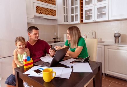 一家人年轻的父母和女儿幸福而美丽，在家里一起微笑，在厨房里用笔记本电脑购物