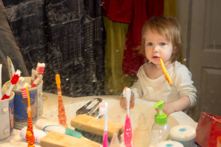 透过肮脏的牙膏飞溅的镜子看一个小女婴。 孩子刷牙，从浴室里的第一颗乳牙上摘下牌匾，那里的一切都乱七八糟。