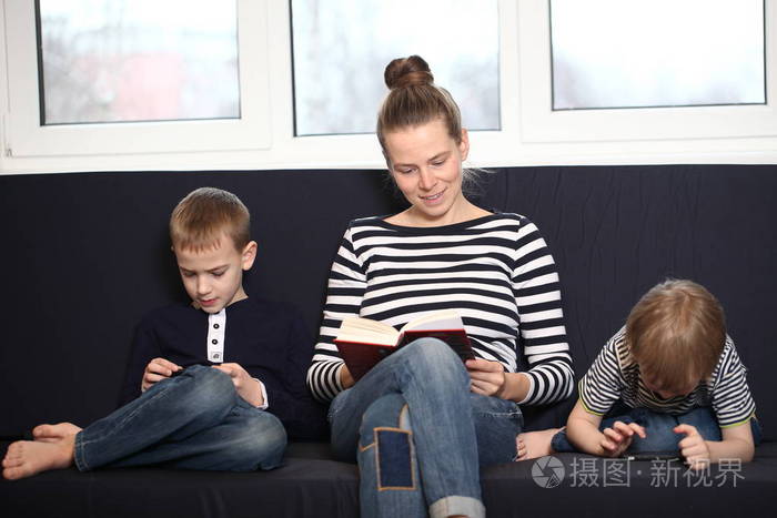 妈妈带着两个兄弟金发男孩在家里的蓝色沙发上玩智能手机