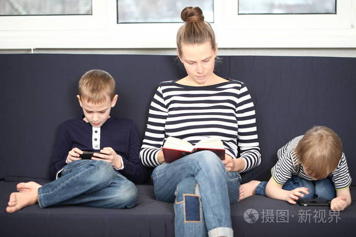 妈妈带着两个兄弟金发男孩在家里的蓝色沙发上玩智能手机