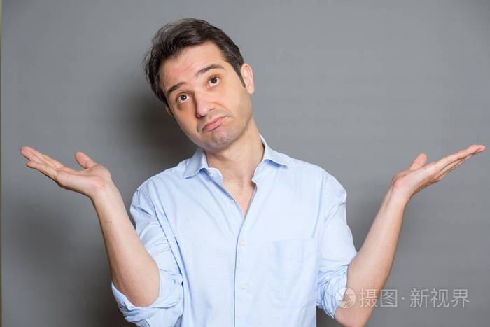 迷糊的男人穿着衬衫站着在灰色的背景上耸耸肩