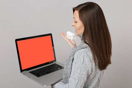 侧视图的妇女在毛衣围巾与纸巾持有笔记本电脑电脑与空白空屏隔离在灰色背景。健康生活方式在线治疗寒冷季节概念