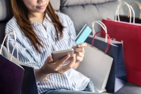 快乐的亚洲女人坐在沙发上，拿着信用卡和智能手机购物，网上支付和五颜六色的购物袋，网上购物的概念