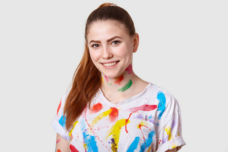快乐快乐的欧洲女艺术家有马尾辫，牙齿微笑，白色，甚至牙齿，穿着休闲T恤，在白色工作室墙上隔离的创造性过程后，被水彩弄脏了。