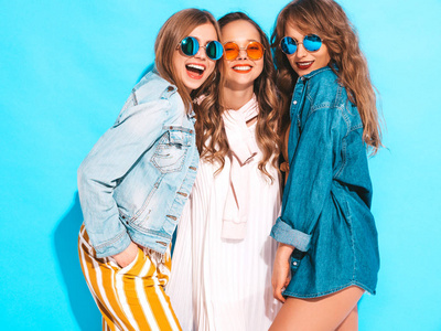 三个年轻美丽的微笑时髦女孩穿着时髦的夏季休闲服装。 无忧无虑的女人在蓝墙附近摆姿势。 积极的模特发疯了