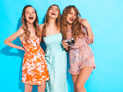 三个年轻美丽的微笑时髦女孩穿着时髦的夏季休闲服装。 无忧无虑的女人在蓝墙附近摆姿势。 拍摄复古相机